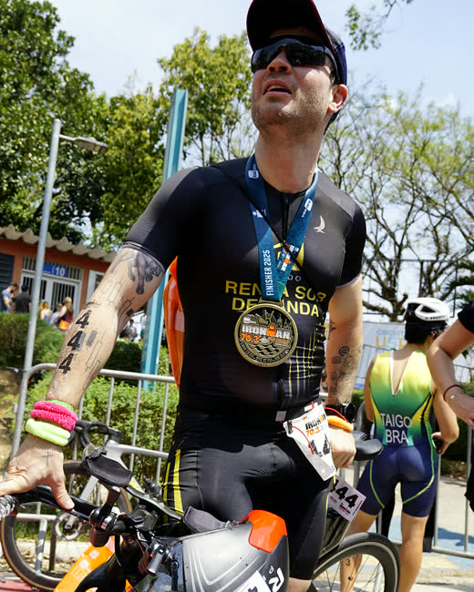 Danilo Zanini com a medalha de finisher do Ironman 70.3 São Paulo 2025, ao lado da bicicleta, com o uniforme escrito Renda Sob Demanda